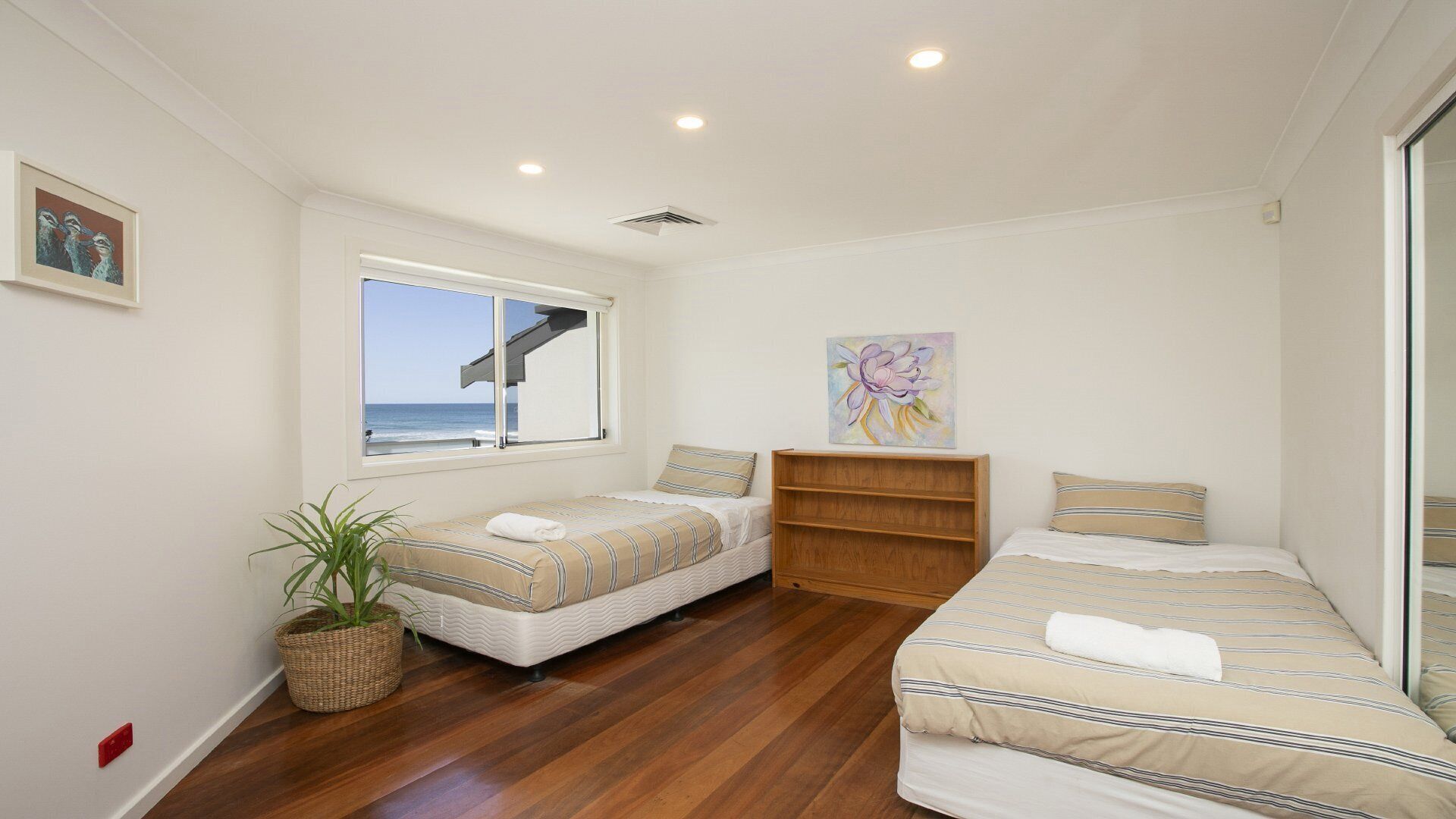A bedroom with two beds, a wooden floor, a window with a view of the sea, a plant, and a shelf with a painting.