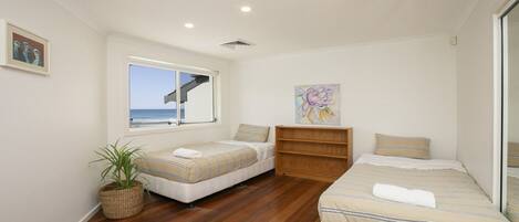 A bedroom with two beds, a wooden floor, a window with a view of the sea, a plant, and a shelf with a painting.