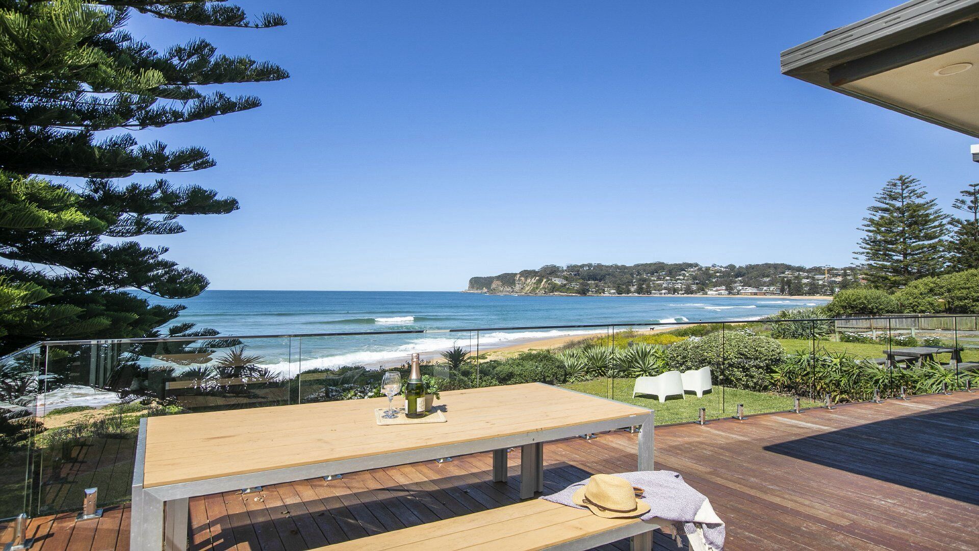 A wooden deck with a table and chairs overlooking a beach with waves and a distant cliff.