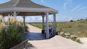 A gazebo with a sloped roof, a concrete pathway leading to it, and a grassy area with tall grasses.