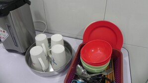 A kitchen area with a stainless steel coffee maker, a tray with white mugs, and a basket containing red and green plates and utensils.