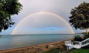Une plage avec une terrasse et des chaises blanches, un arc-en-ciel se déployant au-dessus de l’eau, et des arbres encadrant le paysage.