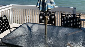Ett bord med glasskiva och metallram, ett randigt parasoll och svarta stolar med ett flätat ryggstöd, med utsikt över en strand.