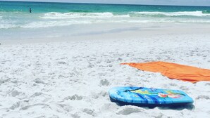 Ein Strand mit weißem Sand, einem blauen aufblasbaren Brett und einem orangefarbenen Handtuch.