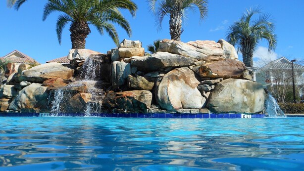 Una piscina con cascada de roca, rodeada de palmeras y un cielo azul despejado.