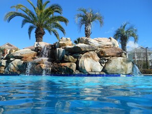 Une piscine agrémentée d’une cascade artificielle en roches, entourée de palmiers et surplombée d’un ciel bleu azur.