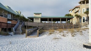 Una propiedad frente a la playa con un paseo marítimo de madera que conduce a un área cubierta, una escalera y edificios con techos verdes.