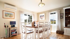 A dining area with a wooden table and white chairs, a television on a side table, and a bookshelf with decorative items. - BlueMill Cottage Bright - The Complete Luxury Getaway (Bright)