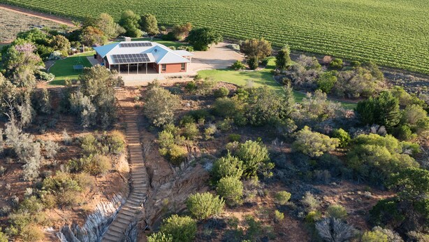 An aerial view of a property with a large house featuring a solar panel roof, surrounded by a vineyard and a natural landscape with a stone pathway leading to the house. - Pet friendly accommodation on the Murray River (Murtho)