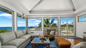 Ein geräumiges Wohnzimmer mit einer großen Couch, einem Glastisch und einem farbenfrohen Sessel. Große Fenster bieten Blick auf das Meer und Palmen.