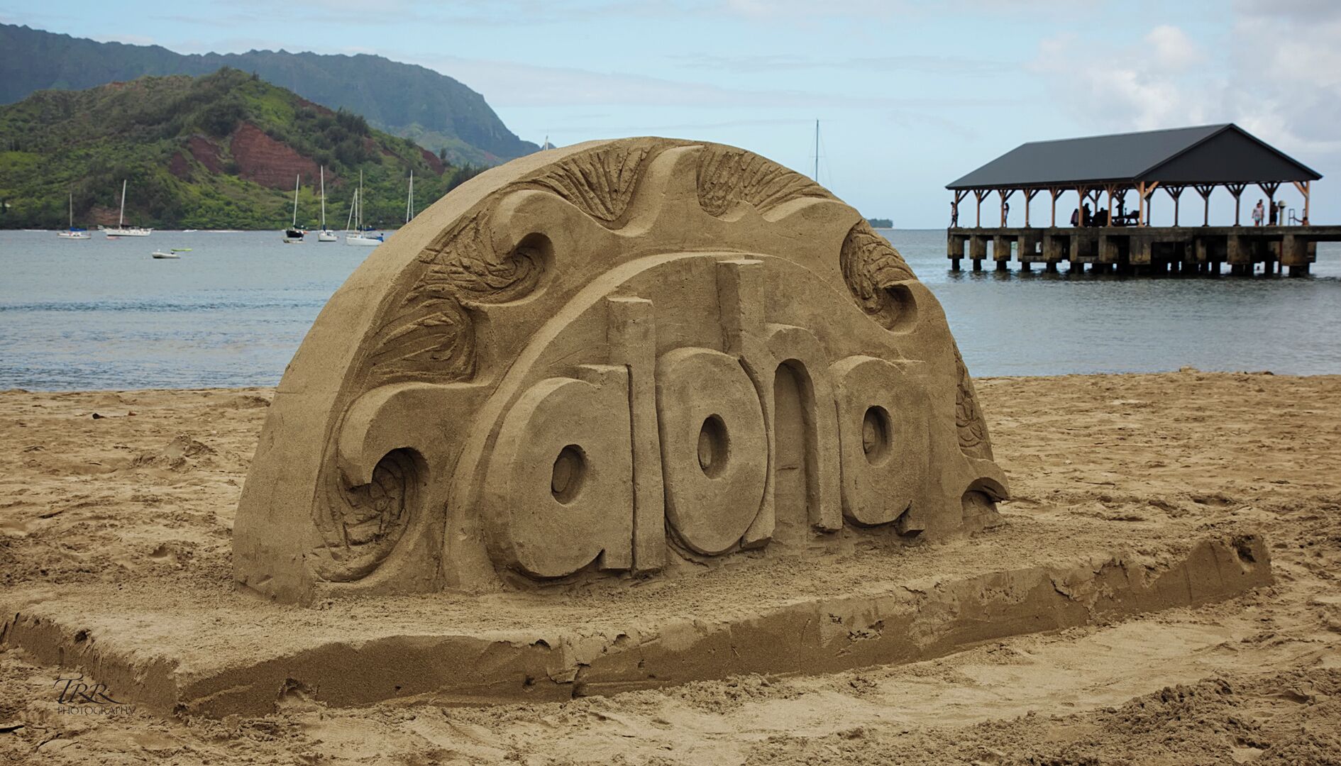 A sand sculpture spelling "Gabriela" on a beach with a pier and boats in the background.