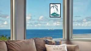 Ein Zimmer mit einem großen Fenster mit Meerblick, ein braunes Sofa mit einem Kissen mit der Aufschrift 'Das Leben ist leicht mit einer Meeresbrise' und ein gerahmtes Delphin-Logo am Fenster.