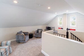 Een loft-achtige kamer met een hoge plafond, twee ramen met witte kozijnen, een trap met zwarte leuningen, en een mix van moderne en klassieke meubels.