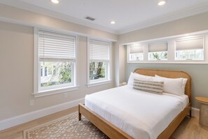 Un dormitorio con una cama grande, tres ventanas con persianas, una mesita redonda y una rejilla en el techo.