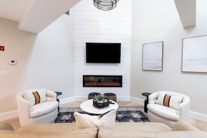 Een moderne woonkamer met een open haard, twee fauteuils, een bank en een flatscreen televisie aan de wand.