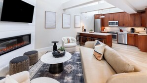 A modern living room with a fireplace, sofa, coffee table, and kitchen area with wooden cabinets and appliances. - 2 Bedrooms w/Loft  and  En-Suite Baths, Walk to Harbor (Newport)