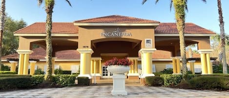Entrada de hotel con pavimento a cuadros, palmeras y una maceta grande con flores.