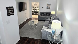 Uma sala de estar moderna com um sofá cinza, uma mesa de centro de vidro e uma TV fixada na parede.