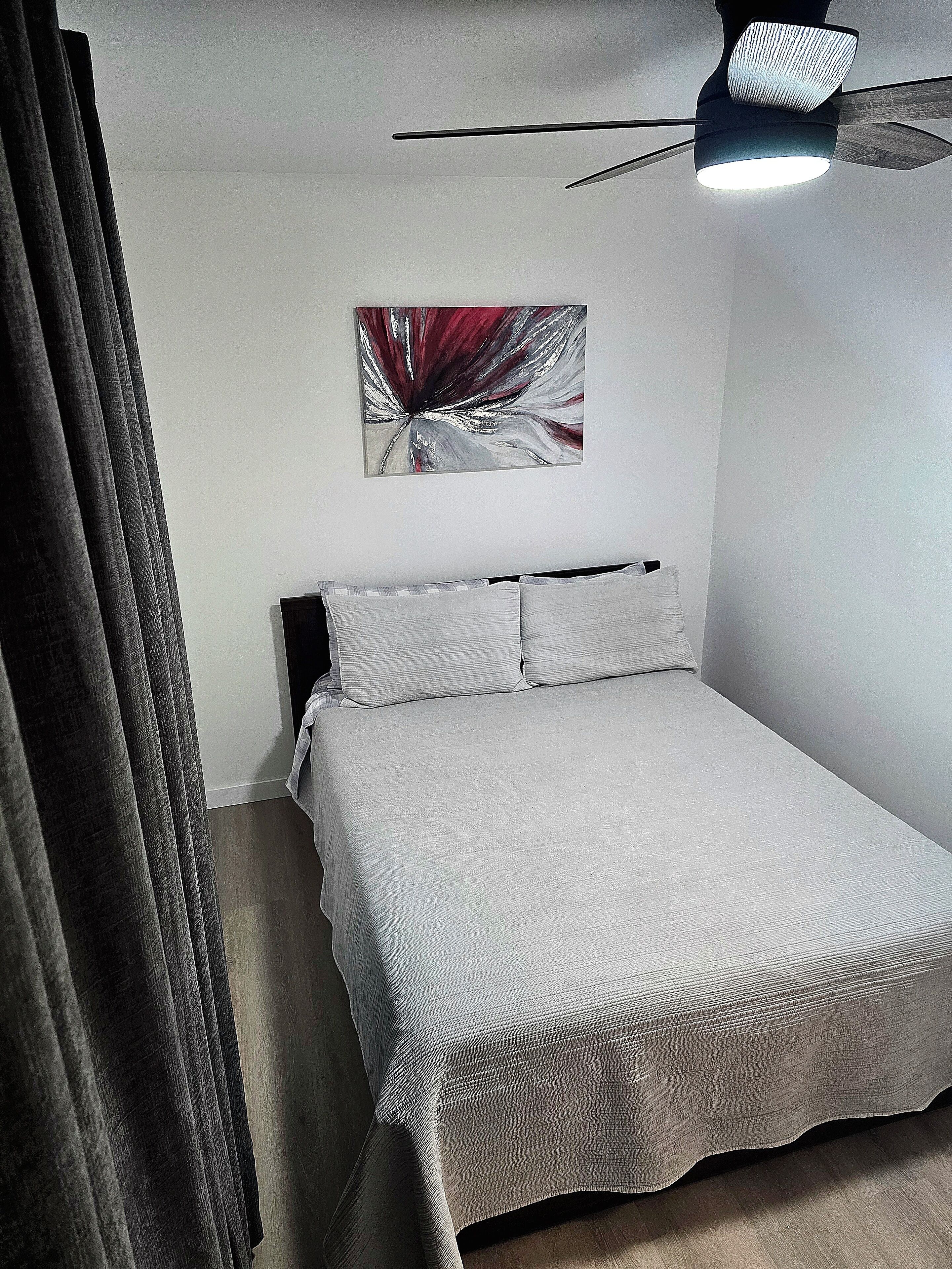 A bedroom with a bed, a ceiling fan, and a wall-mounted abstract painting.