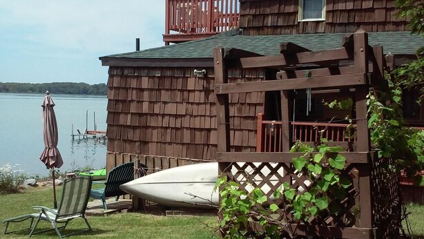Eine Holzhütte mit einer Terrasse mit Blick auf einen See, einem Boot und einem Stuhl am Wasser.