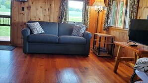 Ein gemütliches Blockhaus-Interieur mit Holzbalkendecke, einer Couch mit gemusterten Kissen, einem Fernseher auf einem Holztisch und einem Fenster mit Vorhängen.