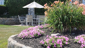A garden with pink flowers, a stone bench, and a table with chairs under a white umbrella. - Exclusive Private Beach Neighborhood offers Spectacular Summer Home (Narragansett)