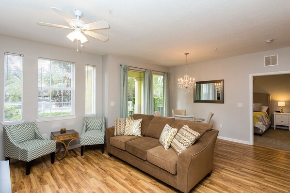 A living room with a brown sofa, patterned cushions, a wooden floor, a ceiling fan, and large windows with blinds. - Newly Renovated, Modern Two Bedroom Condo Minutes Away From Disney (Kissimmee)