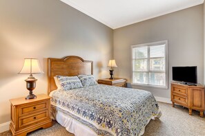 Une chambre avec un grand lit, deux tables de chevet avec des lampes, un téléviseur posé sur un meuble en bois et une fenêtre avec des stores.
