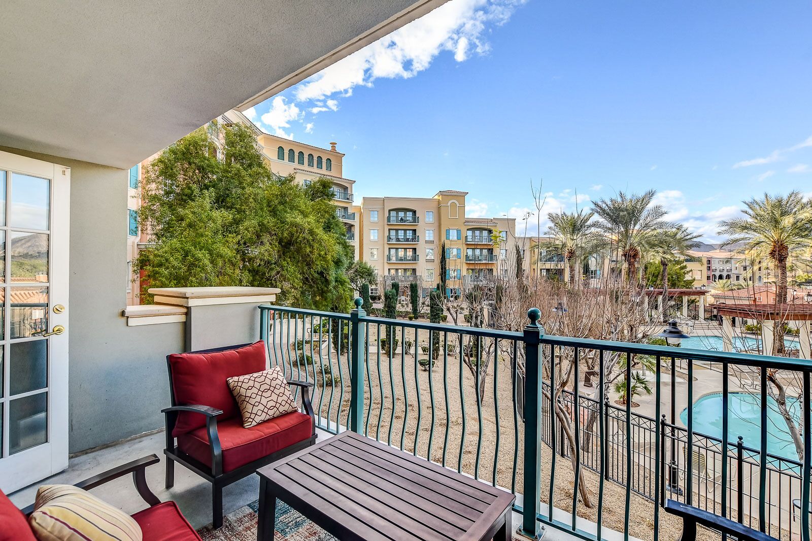 Een balkon met een rode stoel en een houten tafel, met uitzicht op een zwembad en een woestijnlandschap met gebouwen op de achtergrond.