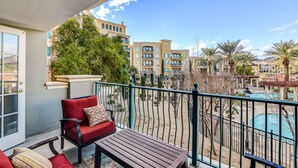 A balcony with a red chair and a wooden table, overlooking a pool and a desert landscape with buildings in the background.