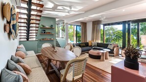 A modern living room with a dining area, a staircase, and large windows offering a view of greenery. - Point Blue, Beautiful House on Hamilton Island (Hamilton Island)