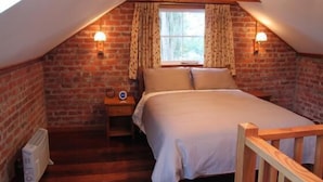A cozy attic bedroom with a wooden bed, two bedside lamps, a window with curtains, and a radiator. - Historic brick stables (Otaki)