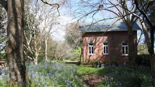 A red brick house with a pathway leading to it, surrounded by trees and purple flowers. - Historic brick stables (Otaki)