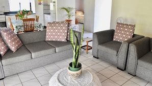 Ein Wohnzimmer mit grauem Sofa, gemusterten Kissen, einer Zimmerpflanze in einem Topf und einem Essbereich mit Tisch und Stühlen.