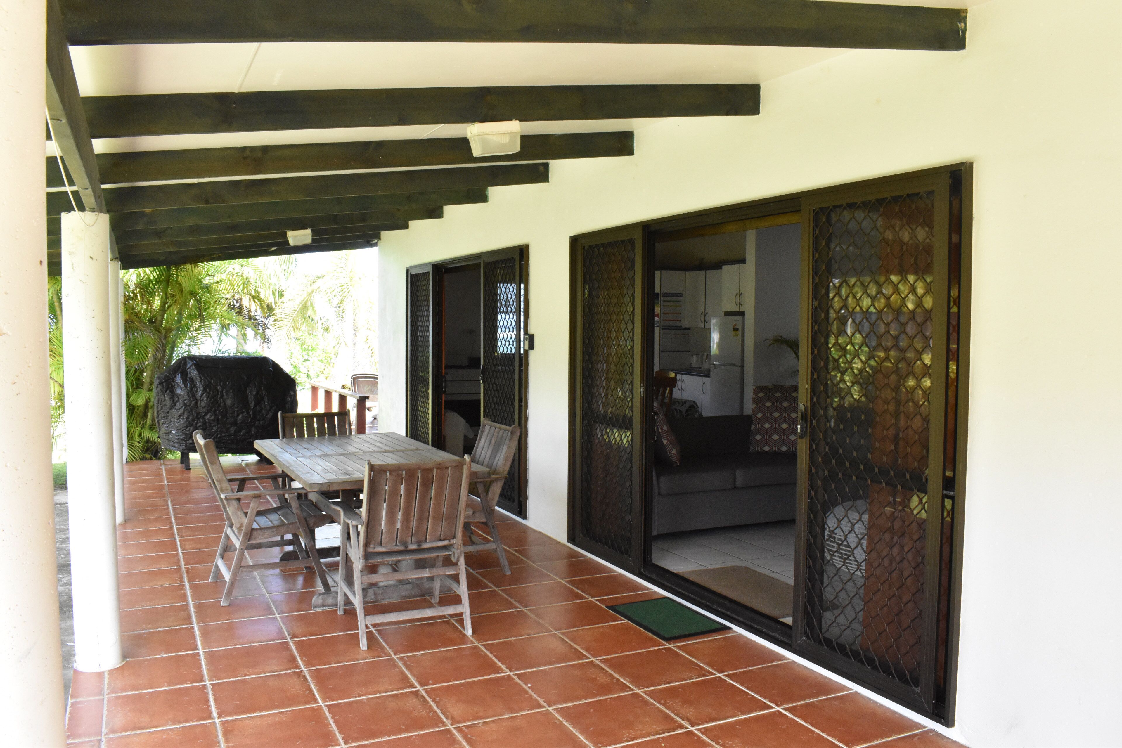 Um pátio externo com mesa e cadeiras de madeira, churrasqueira e porta de vidro deslizante que dá para uma sala de estar.