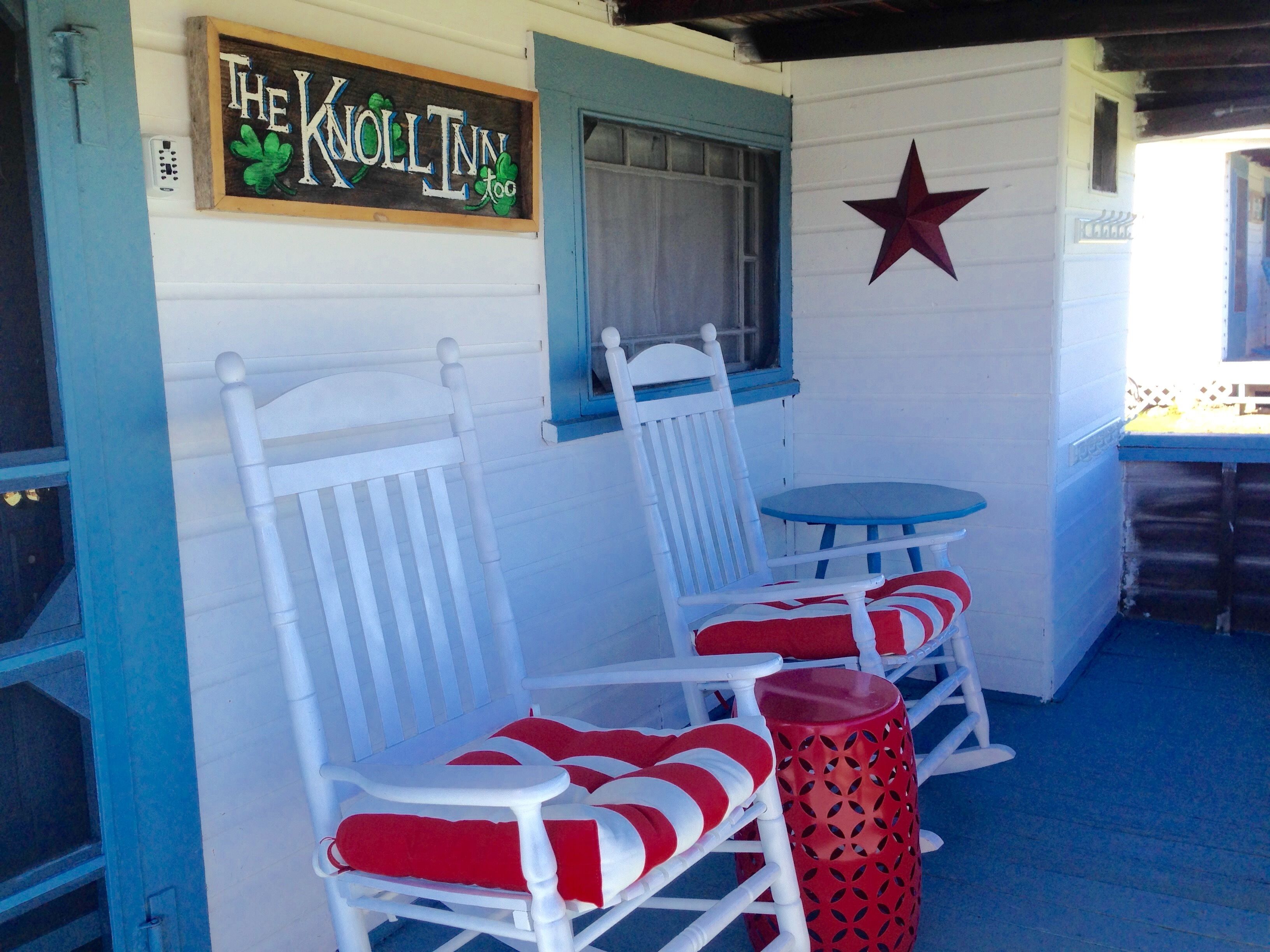 A porch with two white rocking chairs and a small table, a sign reading "The Knoll Inn too", and a red star decoration on the wall.
