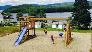 A playground with a slide and swings overlooking a lake with houses and mountains in the background.