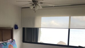 Habitación de hotel con cama, escritorio, silla, ventilador de techo y un ventanal con vista al mar.