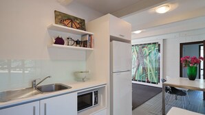 Une cuisine moderne avec des meubles blancs, un évier en acier inoxydable et un four encastré. On y trouve également un réfrigérateur, un four à micro-ondes et une petite étagère où sont disposés des objets décoratifs.