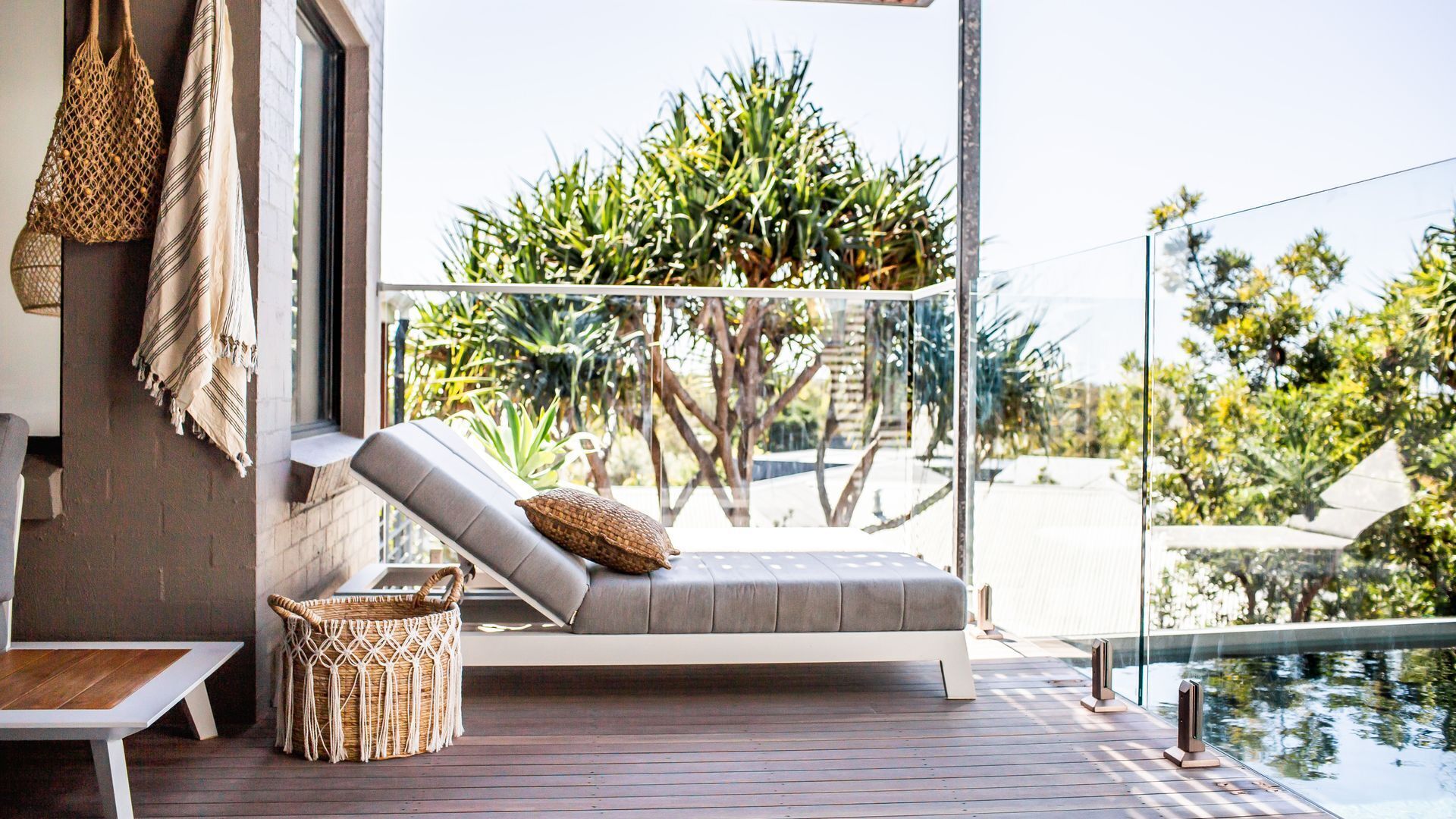 A modern outdoor seating area with a cushioned bench, a woven basket, and a view of tropical plants and a pool.