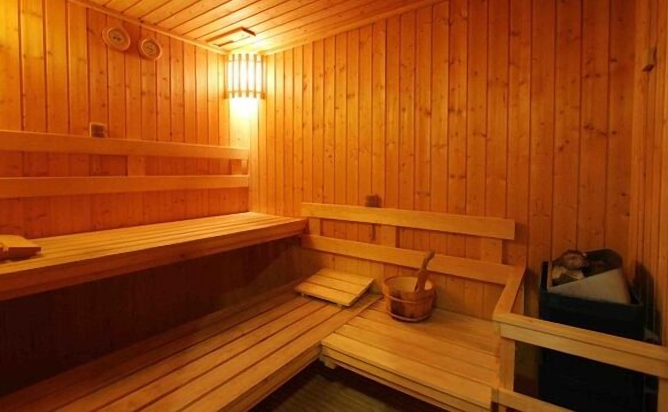 A wooden sauna with benches and a bench seat, a wall-mounted heater, and a bucket with a brush.