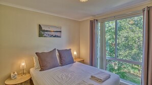 Un dormitorio con una cama grande, dos mesitas de noche, una lámpara de noche, un cuadro en la pared y una ventana con cortinas.