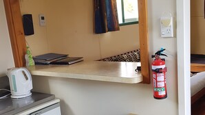 Una cocina compacta con un refrigerador pequeño, una tetera, un estante de madera con una olla de cocción lenta, un extintor de incendios y un reloj en la pared.