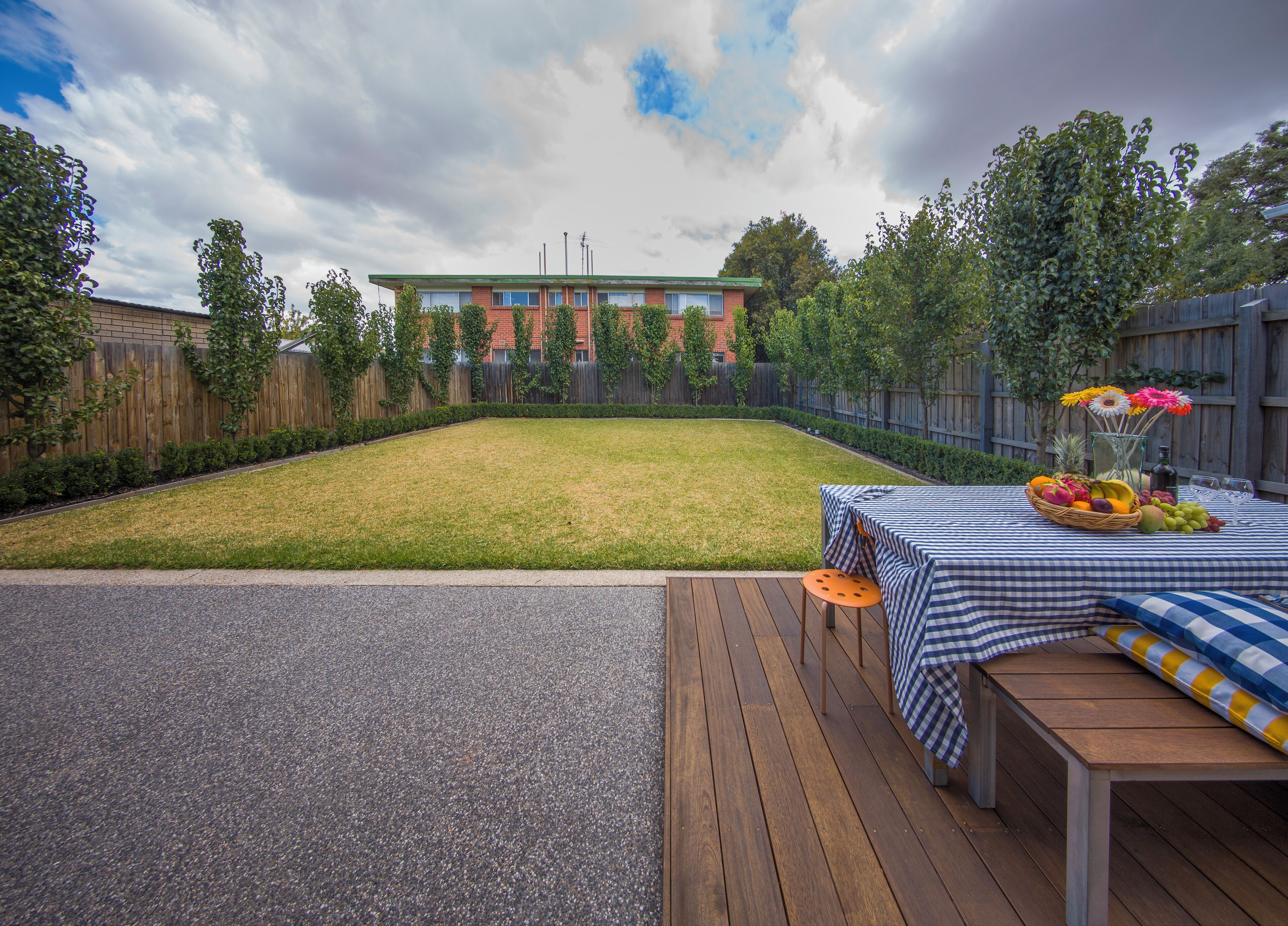 A backyard with a wooden deck, a table with a checkered tablecloth, and a fruit basket.