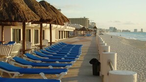 A beachside hotel with rows of blue lounge chairs, thatched-roof structures, and a view of the ocean.