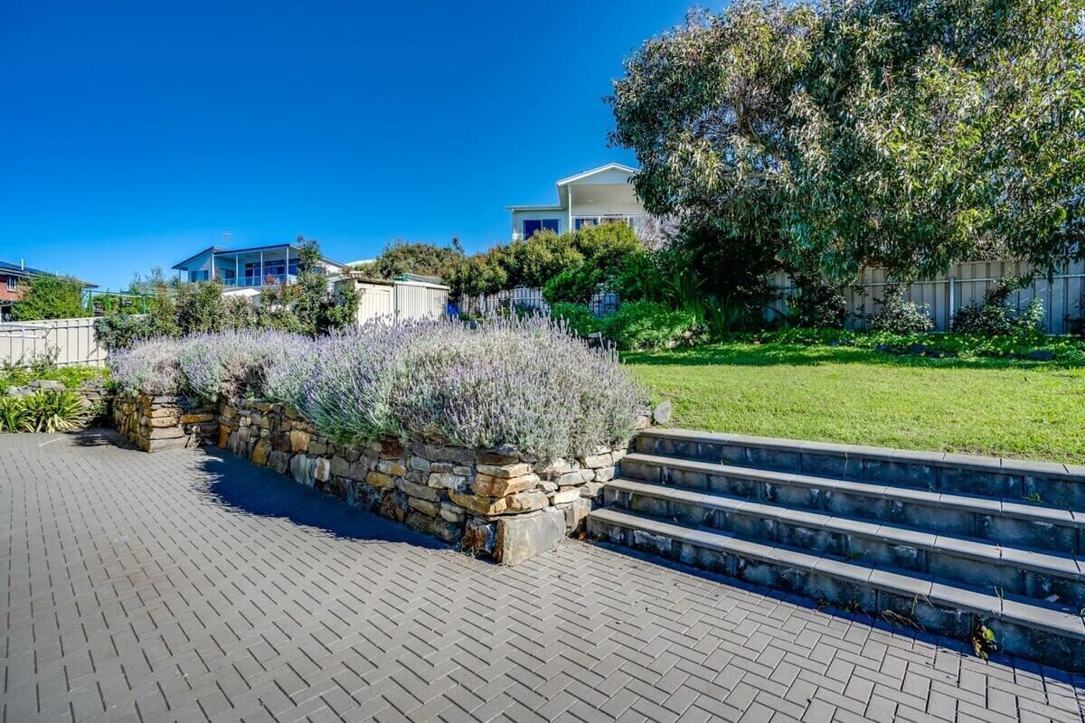 A paved pathway leading to a stone wall with lavender plants, a tree-lined grassy area, and a building with a white facade in the background.