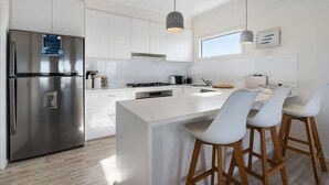 Una cocina moderna con armarios blancos, un refrigerador de acero inoxidable, una isla central con taburetes de bar, iluminación colgante y una ventana que proporciona luz natural.