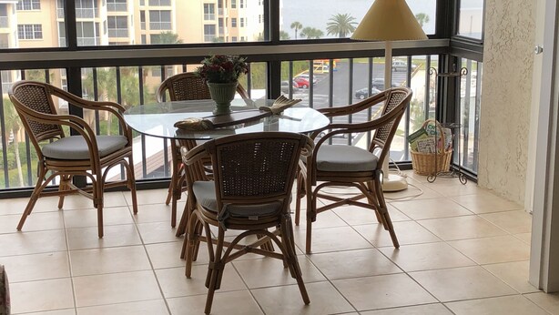Varanda com cadeiras de vime e uma mesa de vidro, com vista para um corpo de água e edifícios.
