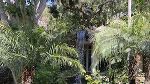 A lush garden with a waterfall, dense foliage, and a stone structure. - Romantic Getaway for 2 in Cottage in complex right on the beach (Oceanside)