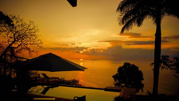 Une vue sur le coucher de soleil depuis un balcon avec une piscine, des palmiers et du mobilier d’extérieur.